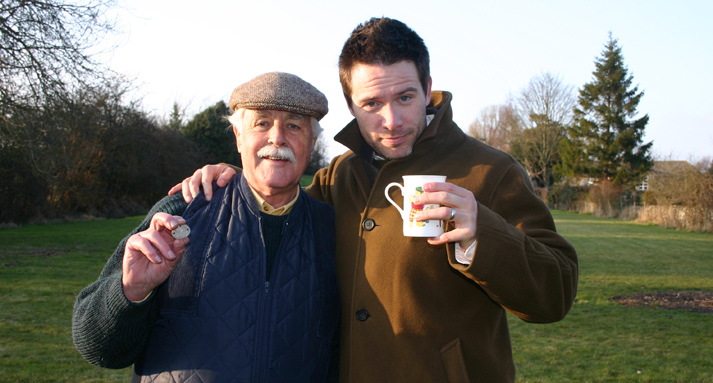 Host Jeff Douglas stands arm-in-arm with an elderly man holding a mug and a small keepsake, in a park.