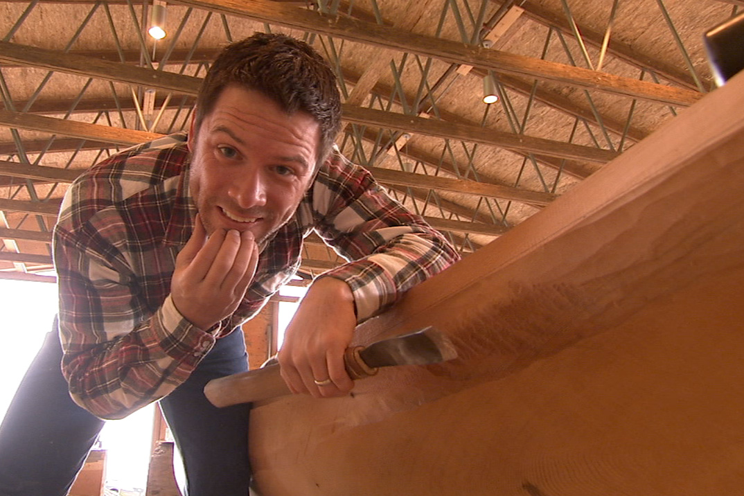 A host shapes a wooden canoe hull with a drawknife inside a timber-framed workshop.