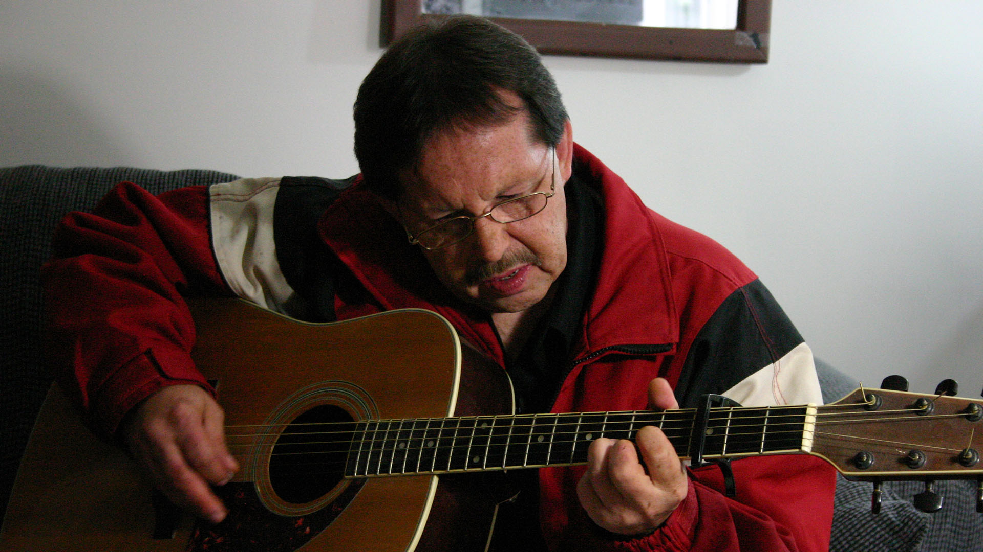 A man plays an acoustic guitar, seated in a living room, one of the film's eight subjects.