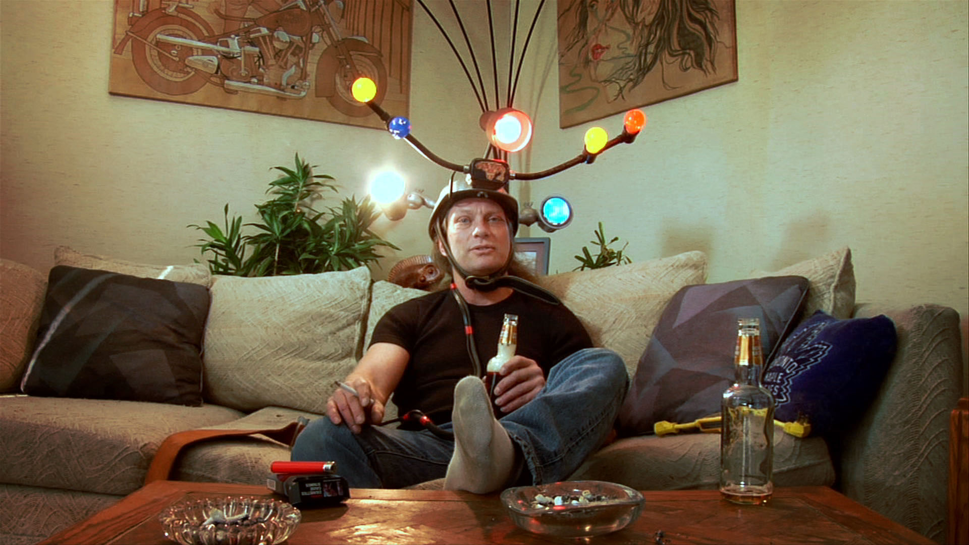 Ralph Zavadil sits on a couch wearing a homemade helmet rigged with colourful lights, holding a beer, surrounded by empty bottles and ashtrays.