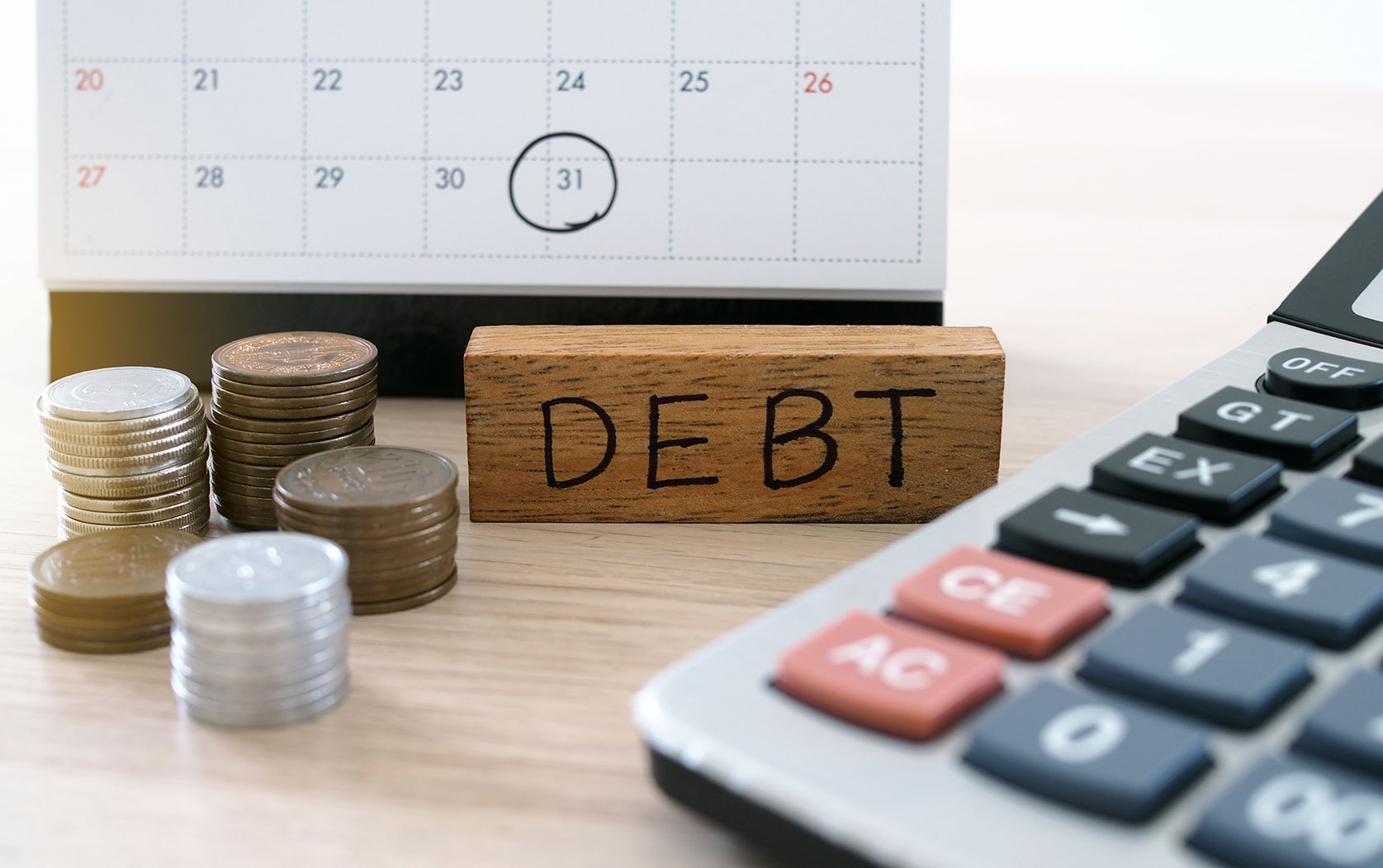 A wooden block reading 'DEBT' sits on a desk beside stacked coins, a calendar and a calculator.