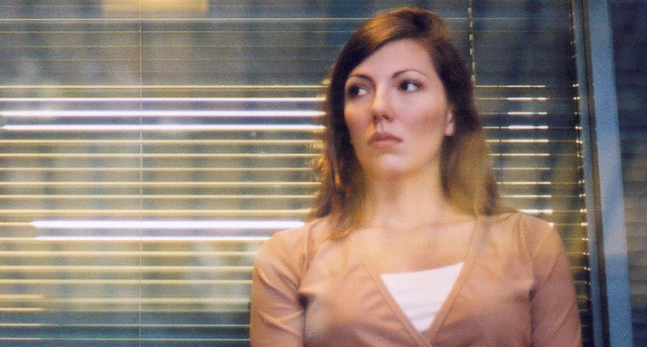A woman glances warily over her shoulder while standing in front of an office window with venetian blinds.