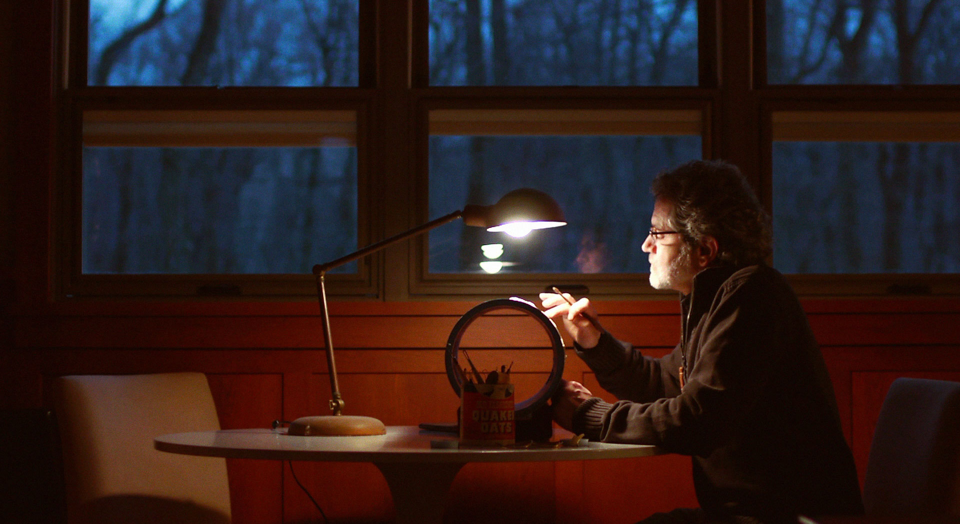 A craftsman leans into a lamplit magnifying glass at a table, working late by a dark, wooded window.
