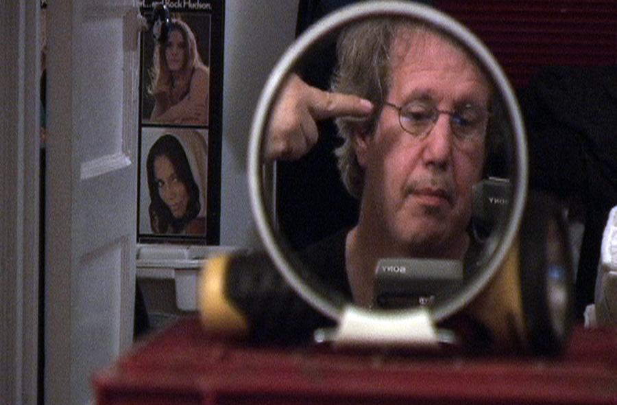 A man examines his reflection in a small round mirror, pointing at his own head, in a cluttered room.
