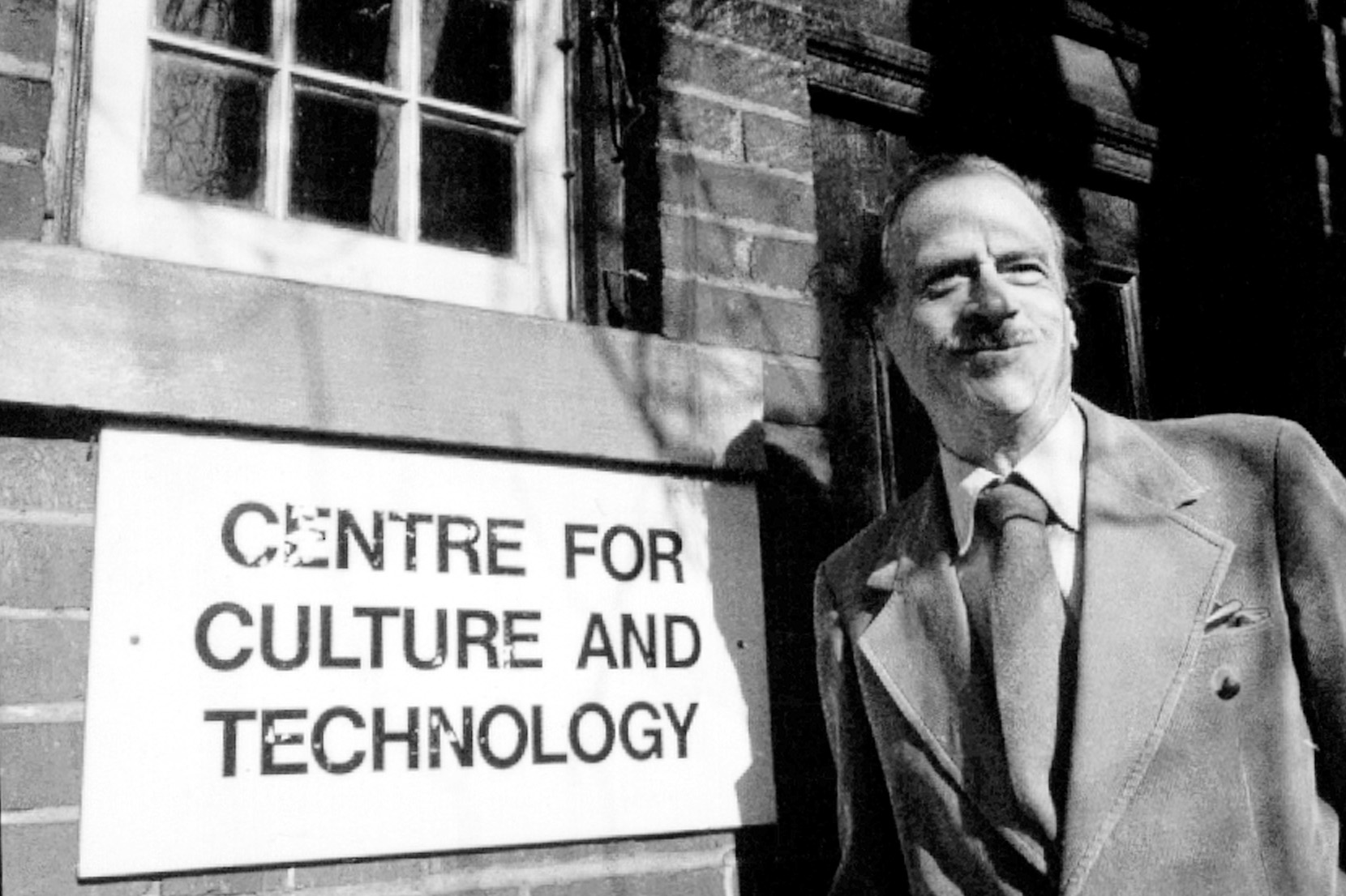Marshall McLuhan stands smiling beside a sign reading 'Centre for Culture and Technology' outside a brick building.