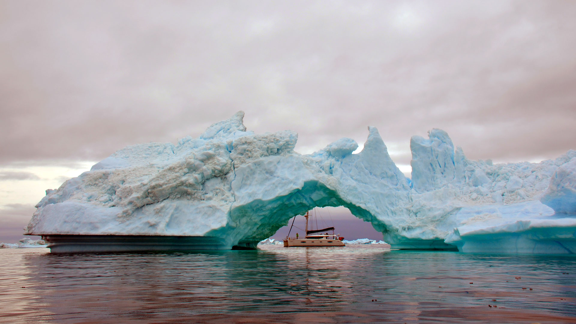 A sailboat passes beneath a massive natural arch carved through a turquoise iceberg.