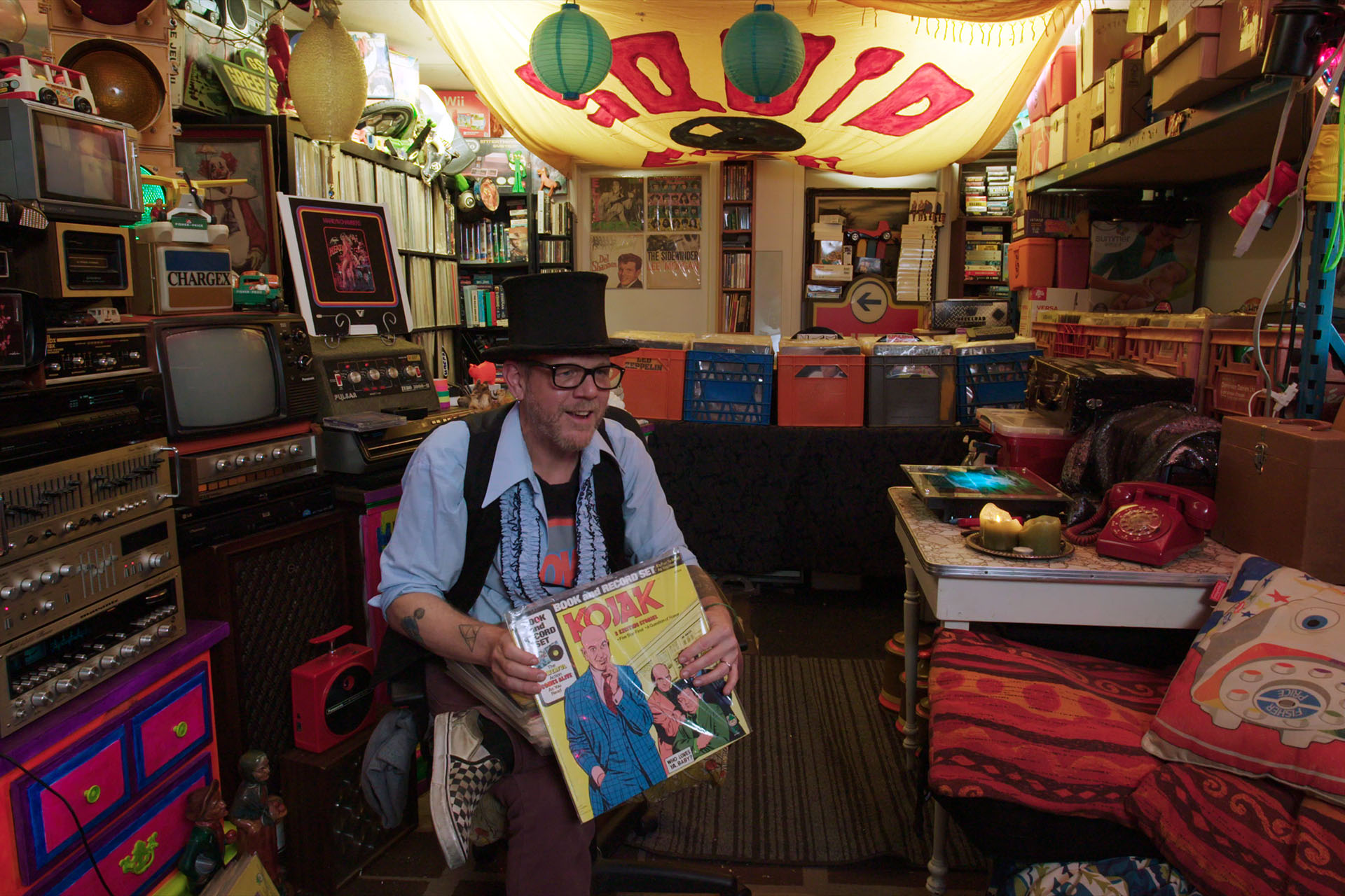 A record collector in a top hat holds up a Kojak record album, surrounded by shelves of vinyl and vintage electronics.