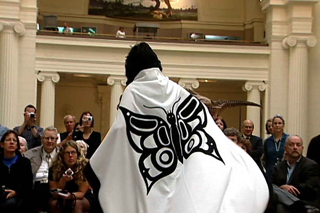 A figure wearing a white button blanket with a black butterfly crest stands before a crowd inside a grand museum hall.