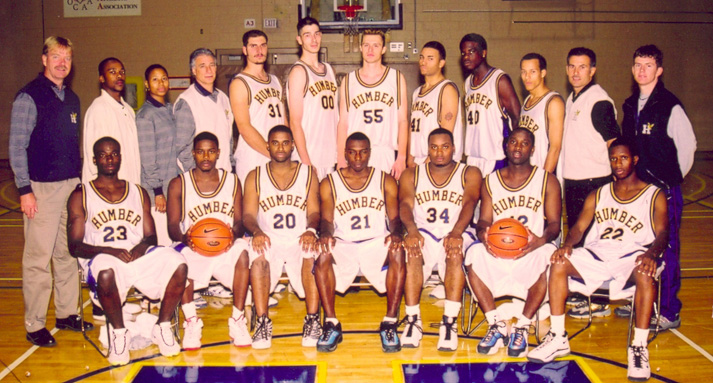 A college basketball team and coaching staff pose together for a team photo in their gym.
