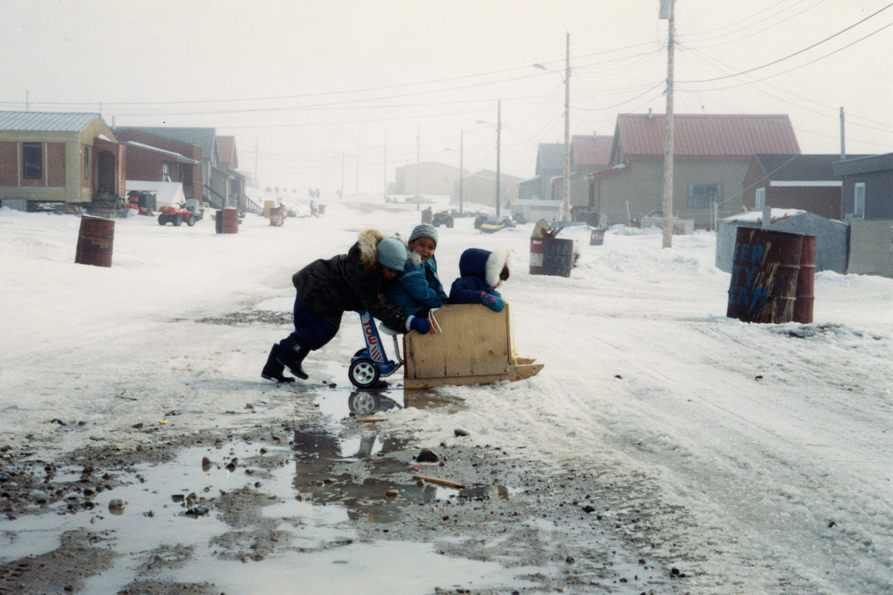 A child pushes two younger children on a homemade wooden sled down a slushy street in an Arctic community.