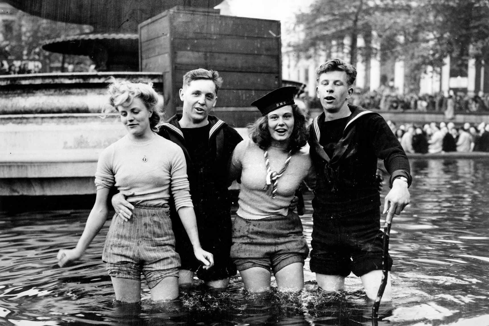 Two sailors and two women wade arm-in-arm through a fountain, celebrating, in a historical black-and-white photograph.