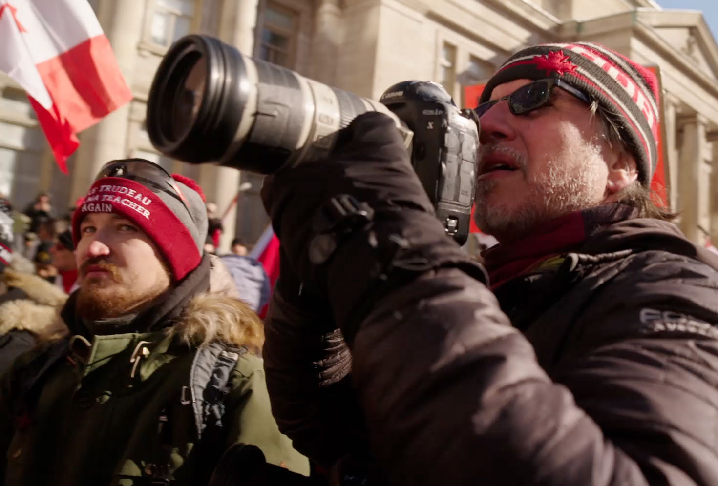 A photographer with a telephoto lens shoots a crowd scene at an outdoor protest, another bystander beside him.