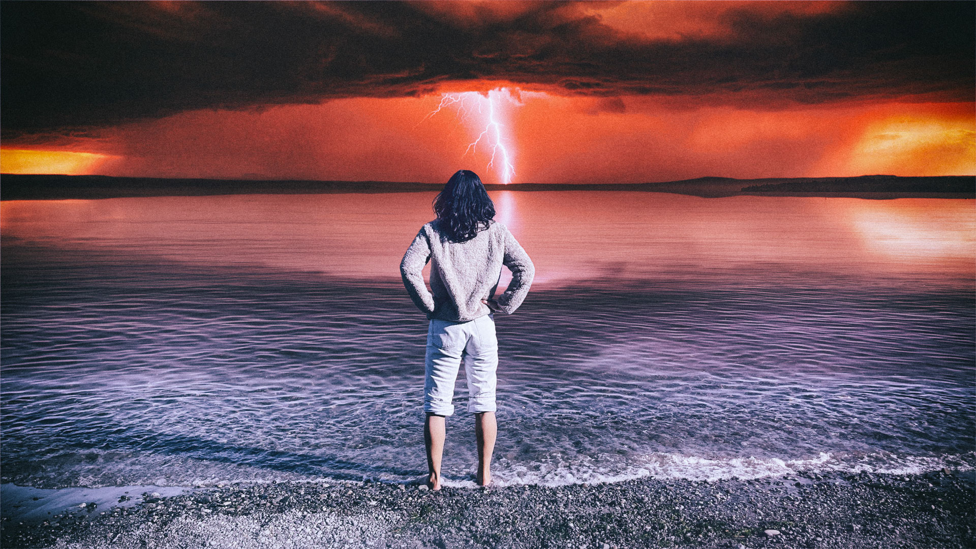 A woman stands on a beach facing a dramatic lightning storm over the water at sunset.